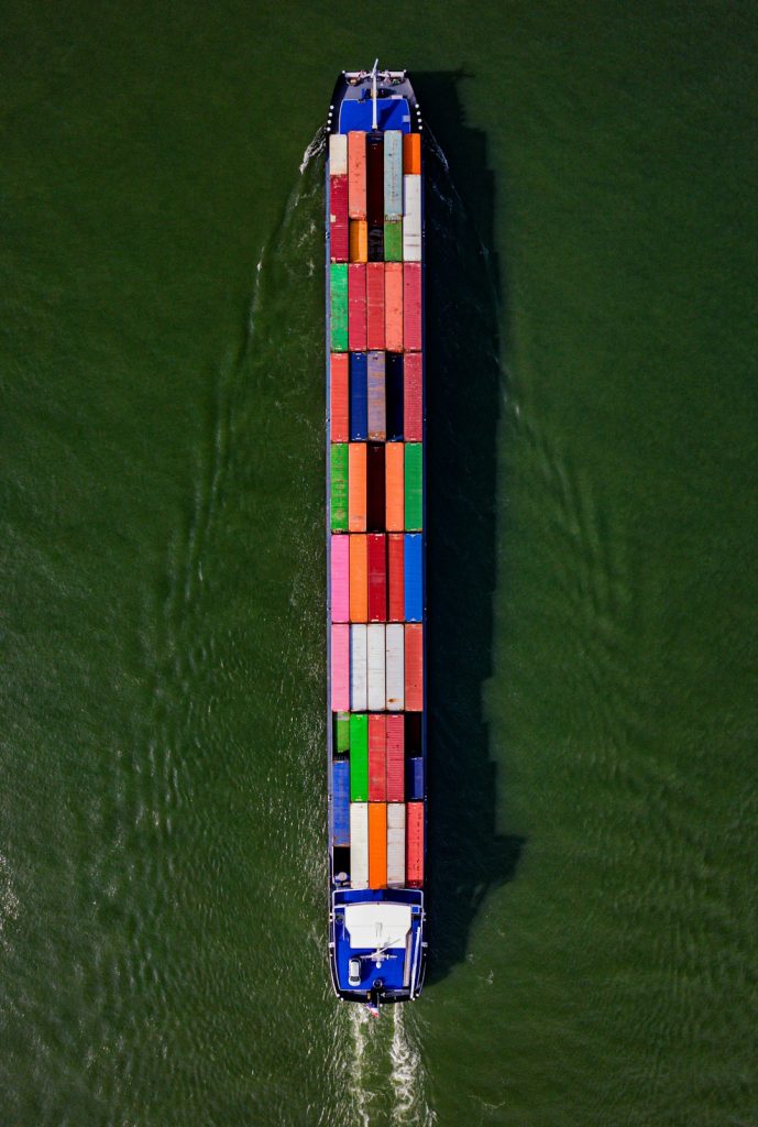 Captured from above, a cargo barge adorned with a mosaic of colorful containers makes its way down a river, vivid against the waters surface, exemplifying the essential role of inland waterways in global trade and logistics.